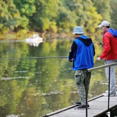 Dwóch mężczyzn łowi ryby z drewnianego pomostu na tle zielonego lasu i spokojnej wody.
