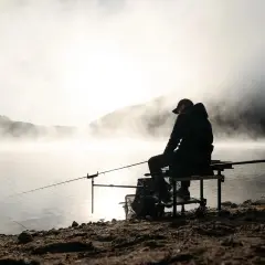 Wędkarz siedzi nad mglistym jeziorem z wędką.