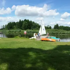 Słoneczny dzień nad jeziorem Kozienickim. Ludzie odpoczywają na trawie, opalają się, a na wodzie pływa żaglówka.
