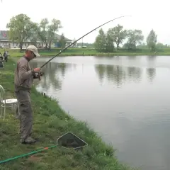 Mężczyzna łowi ryby nad jeziorem GIM. W tle budynek i drzewa.