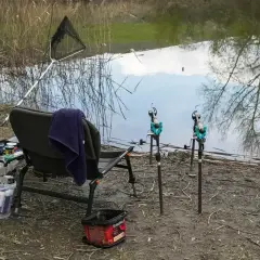 Wędka, podbierak i krzesło nad wodą. Obok stoi wiaderko z zanętą, gotowe do łowienia.