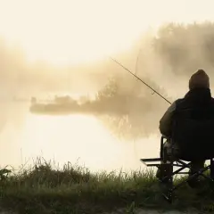 Wędkarz siedzi na krześle nad mglistym jeziorem o poranku. Czeka na branie, ciesząc się spokojem.