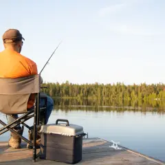 Mężczyzna w pomarańczowej koszulce łowi ryby na pomoście nad jeziorem.
