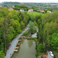 Widok z lotu ptaka na łowisko pstrągowe otoczone zielonymi drzewami, z domami w tle.