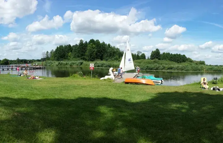 Słoneczny dzień nad jeziorem Kozienickim. Ludzie odpoczywają na trawie, opalają się, a na wodzie pływa żaglówka.