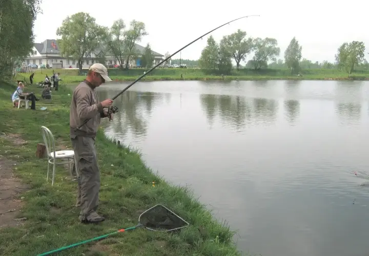 Mężczyzna łowi ryby nad jeziorem GIM. W tle budynek i drzewa.
