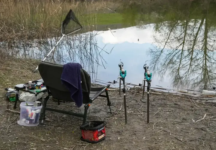 Wędka, podbierak i krzesło nad wodą. Obok stoi wiaderko z zanętą, gotowe do łowienia.