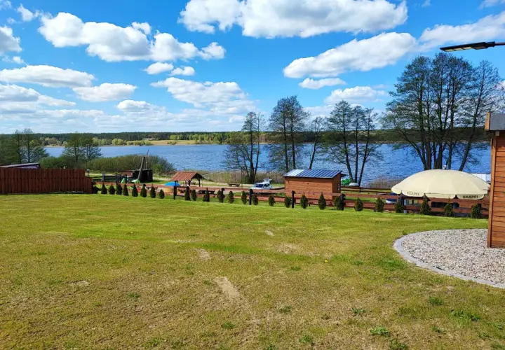 Piękny dzień nad jeziorem skarlińskim. Błękitne niebo z białymi chmurami, zielona trawa i spokojna tafla wody.