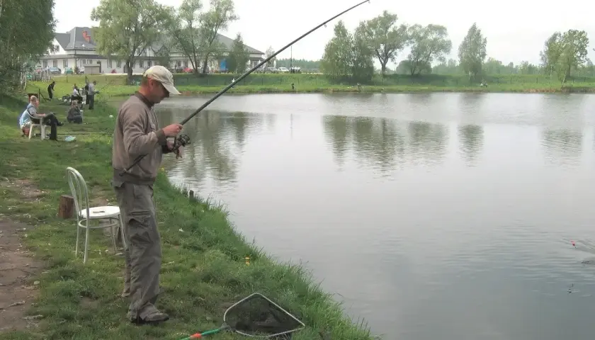 Mężczyzna łowi ryby nad jeziorem GIM. W tle budynek i drzewa.