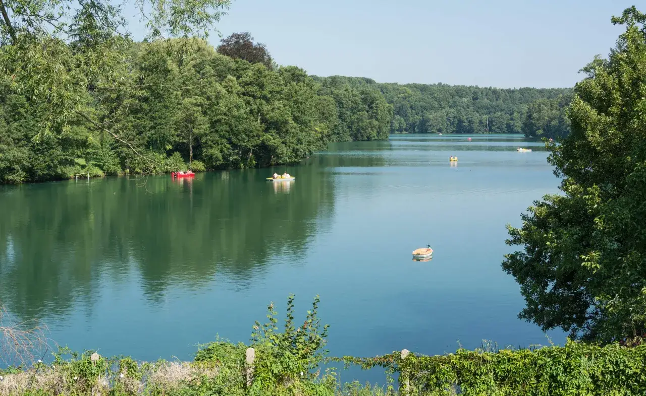 Spokojne jezioro, jego błękitna ciecz odbija zielone drzewa. Kilka łódek pływa po wodzie, tworząc sielski obraz.