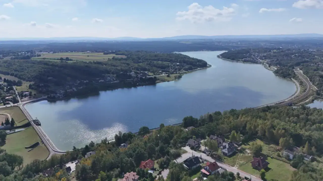Widok z lotu ptaka na zalew Brody otoczony lasami i domami. W oddali widać pola i wzgórza.
